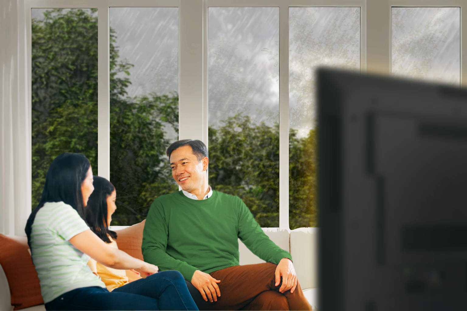 A Filipino family relaxing in their living room with the lights and TV on while heavy rain falls outside the windows